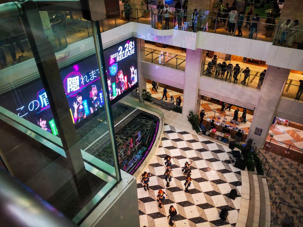 Blick von oben auf ein sonnenbeschienenes Einkaufszentrum mit einer Tanzgruppe, die auf einem schwarz-weiß gekachelten Boden auftritt. Die Kunden schauen von mehreren Stockwerken in der Sunshine City aus zu, während die Sonne hereinströmt und große digitale Bildschirme im Hintergrund japanische Werbung zeigen.
