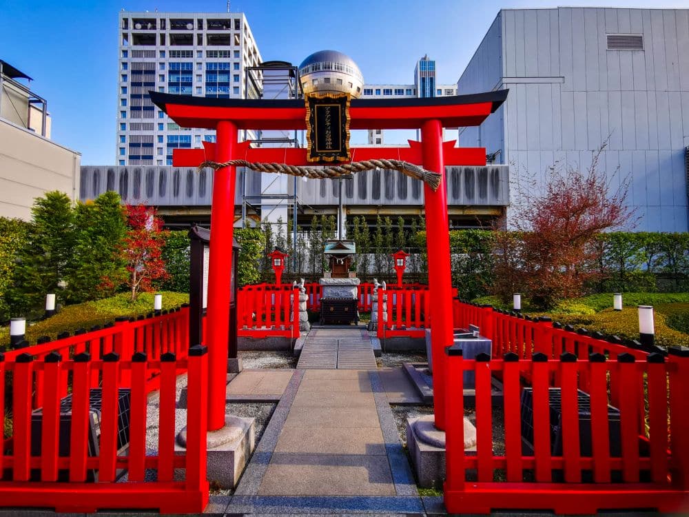 Ein leuchtend rotes Torii-Tor steht am Eingang eines kleinen städtischen Schreins in Odaiba, umgeben von roten Zäunen, Grünpflanzen und modernen Gebäuden im Hintergrund unter einem klaren blauen Himmel.