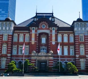 Die Tokyo Station ein historisches Gebäude aus rotem Backstein mit Bogenfenstern und dekorativem Mauerwerk, flankiert von zwei japanischen Flaggen und kleinen Bäumen, steht vor modernen gläsernen Wolkenkratzern unter einem klaren Himmel.