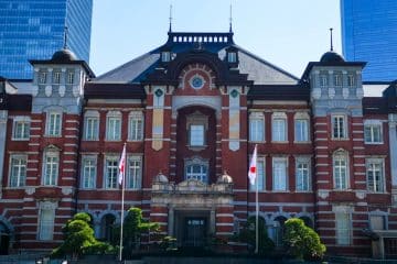 Die Tokyo Station ein historisches Gebäude aus rotem Backstein mit Bogenfenstern und dekorativem Mauerwerk, flankiert von zwei japanischen Flaggen und kleinen Bäumen, steht vor modernen gläsernen Wolkenkratzern unter einem klaren Himmel.