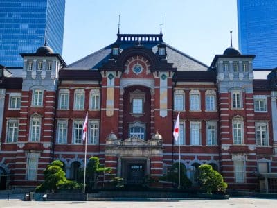 Die Tokyo Station ein historisches Gebäude aus rotem Backstein mit Bogenfenstern und dekorativem Mauerwerk, flankiert von zwei japanischen Flaggen und kleinen Bäumen, steht vor modernen gläsernen Wolkenkratzern unter einem klaren Himmel.