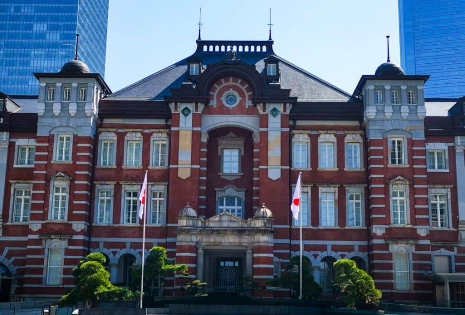 Die Tokyo Station ein historisches Gebäude aus rotem Backstein mit Bogenfenstern und dekorativem Mauerwerk, flankiert von zwei japanischen Flaggen und kleinen Bäumen, steht vor modernen gläsernen Wolkenkratzern unter einem klaren Himmel.