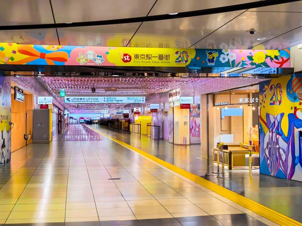Ein farbenfroher, dekorierter Innengang im Bahnhof Tokio mit Schildern, leuchtenden Blumenmustern und Überkopfbannern. Geschäfte säumen den Korridor, während gelbe taktile Pflastersteine auf dem glänzenden Boden auffallen.