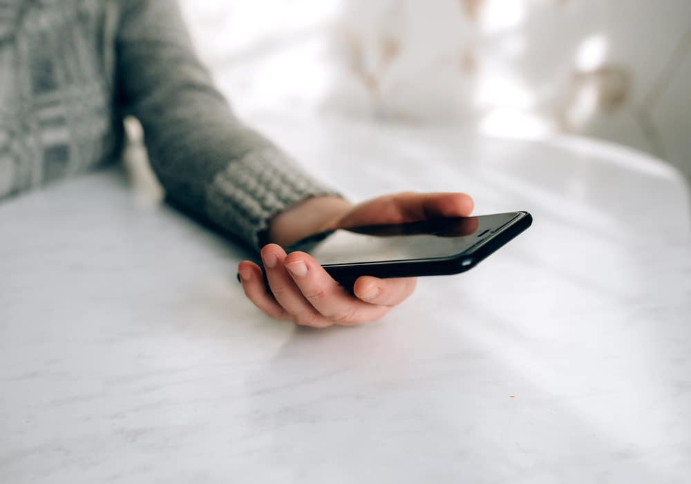 Eine Person in einem grauen Pullover hält ein schwarzes, eSIM-fähiges Smartphone in der rechten Hand und legt es auf einen weißen Marmortisch. Der Bildschirm des Telefons ist nach oben gerichtet, während der Hintergrund leicht unscharf ist.