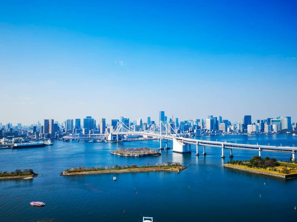 Ein Panoramablick auf die Skyline einer Stadt mit hohen Gebäuden, einer großen weißen Hängebrücke und blauem Wasser im Vordergrund unter einem klaren blauen Himmel. Kleine Boote und grüne Flecken auf dem Wasser in der Nähe von Odaiba.