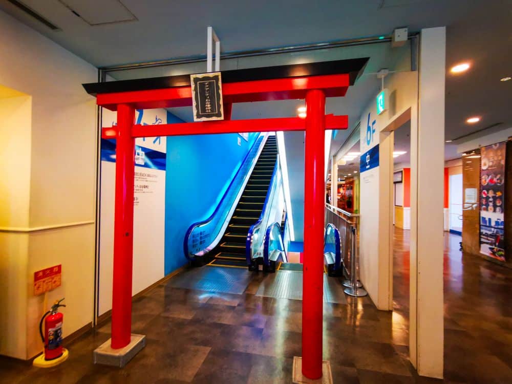 Ein leuchtend rotes Torii-Tor steht im Innenbereich vor einer blau beleuchteten Rolltreppe und verbindet traditionelle japanische Architektur mit dem Ambiente eines modernen Einkaufszentrums oder Unterhaltungszentrums in Odaiba.
