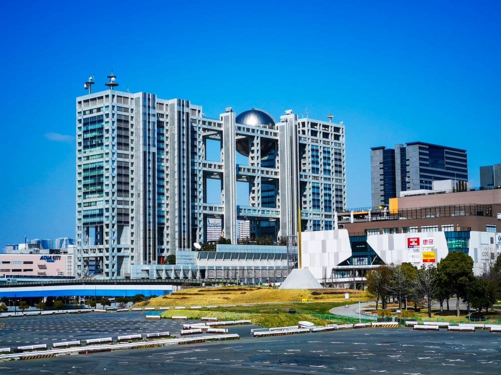 Ein modernes Gebäude in Odaiba, in dessen Mitte eine große silberne Kugel hängt, steht vor einem klaren blauen Himmel, umgeben von anderen städtischen Gebäuden und einem weitgehend leeren Parkplatz im Vordergrund.