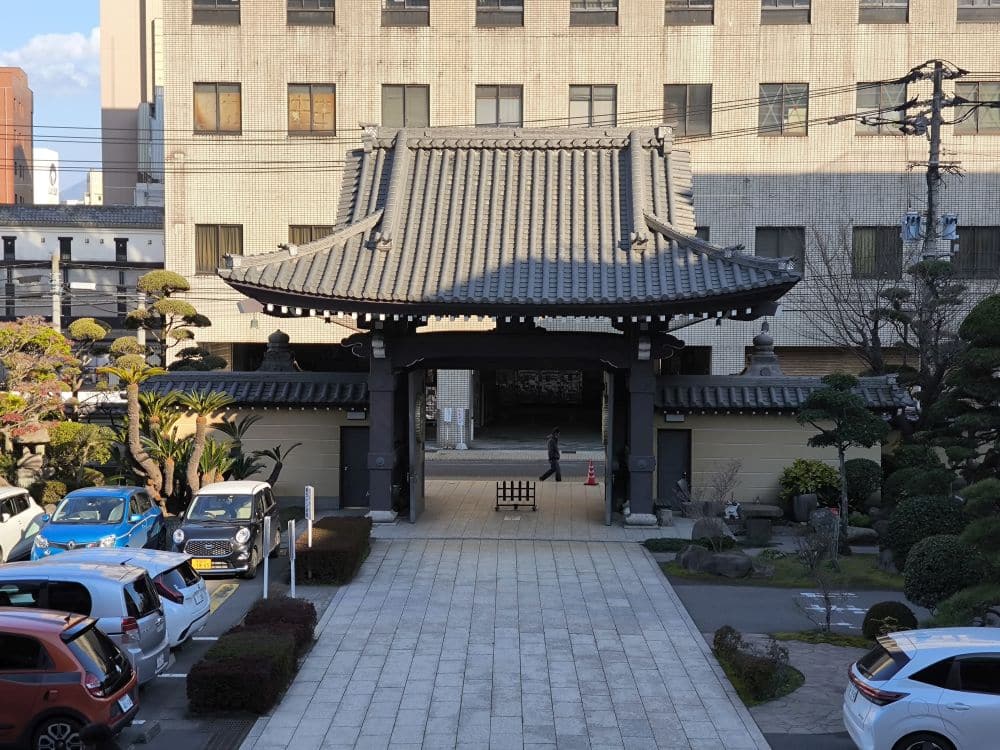 Ein traditionelles Honganji-Tempeltor steht vor einem modernen Gebäude. Ordentlich gestutzte Bäume und geparkte Autos säumen den Steinweg, der zum Eingang führt.