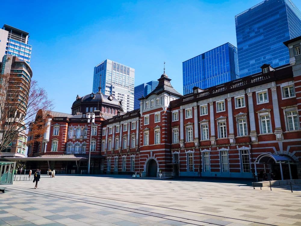 Das historische Gebäude des Tokioter Bahnhofs aus rotem Backstein mit verzierten Fenstern und Dächern steht auf einem weitläufigen Platz, umgeben von modernen gläsernen Wolkenkratzern unter einem klaren blauen Himmel. Nur wenige Menschen schlendern über die Freifläche vor der Tokyo Station.