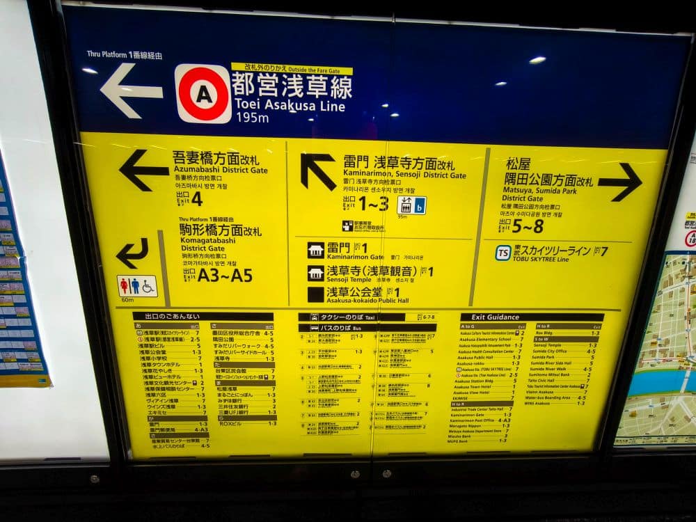 Gelbes Schild der Tokioter U-Bahn für die Toei Asakusa-Linie mit Richtungsangaben, Bahnsteiginformationen, Ausgängen und lokalen Highlights in Japanisch und Englisch, mit einem großen Pfeil nach links und klaren Angaben zum Ausgang.