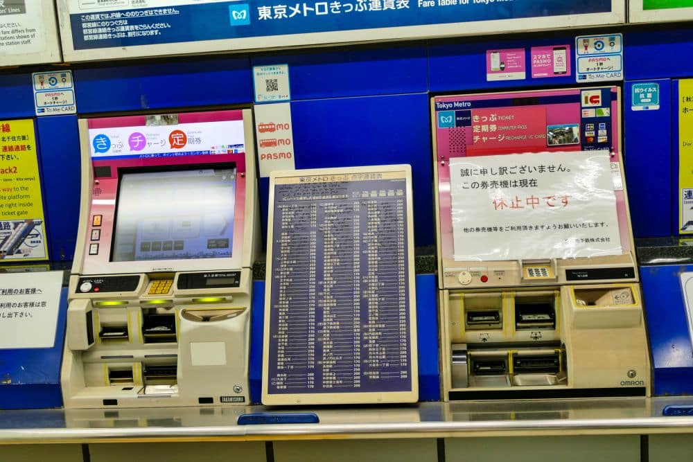 Drei Metro-Fahrkartenautomaten in einer Tokioter Station; zwei sind mit Bildschirmen und Tasten in Betrieb, während der rechte ein außer Betrieb befindliches Schild auf Japanisch zeigt. Über den Automaten sind blaue Informationstafeln zu sehen.