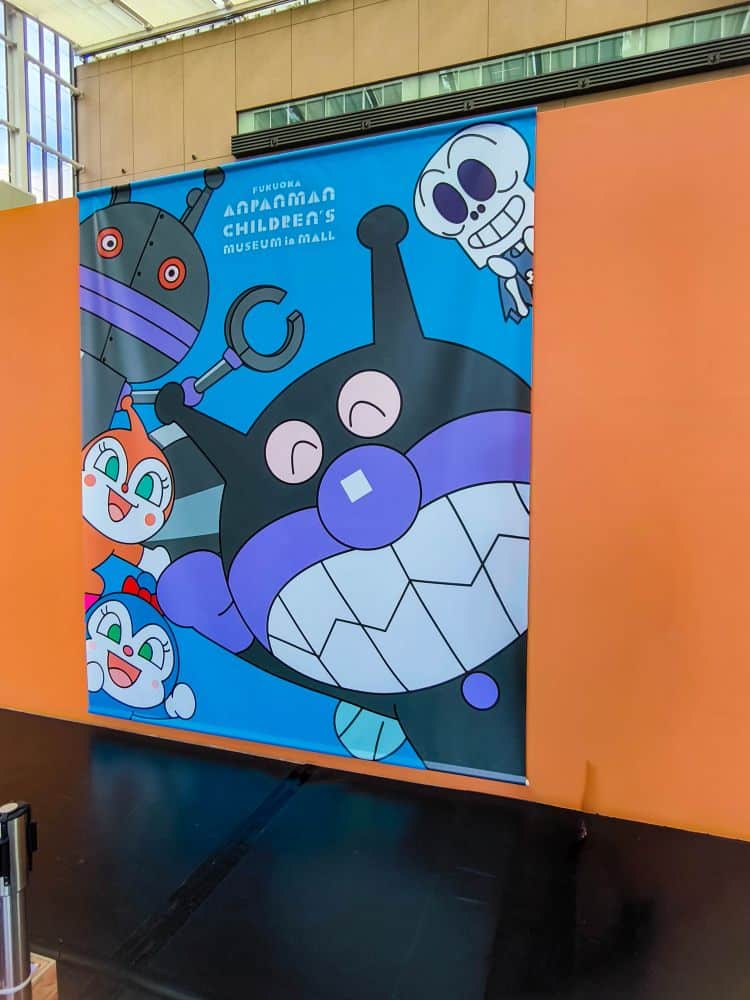 Ein farbenfrohes Anpanman-Banner für das Kindermuseum in einem Einkaufszentrum zeigt Zeichentrickfiguren mit großen Augen und fröhlichen Gesichtsausdrücken auf einem orangefarbenen Hintergrund.