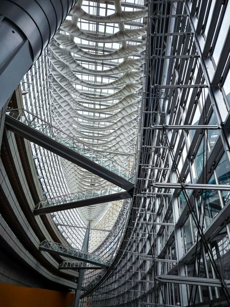 Ein modernes, weitläufiges Atrium aus Glas und Stahl, das vom Tokyo International Forum inspiriert ist, mit einer geschwungenen Decke und einem Fachwerk mit komplizierten geometrischen Mustern und über dem offenen Raum schwebenden Gängen. Durch die großen Fenster strömt natürliches Licht herein.