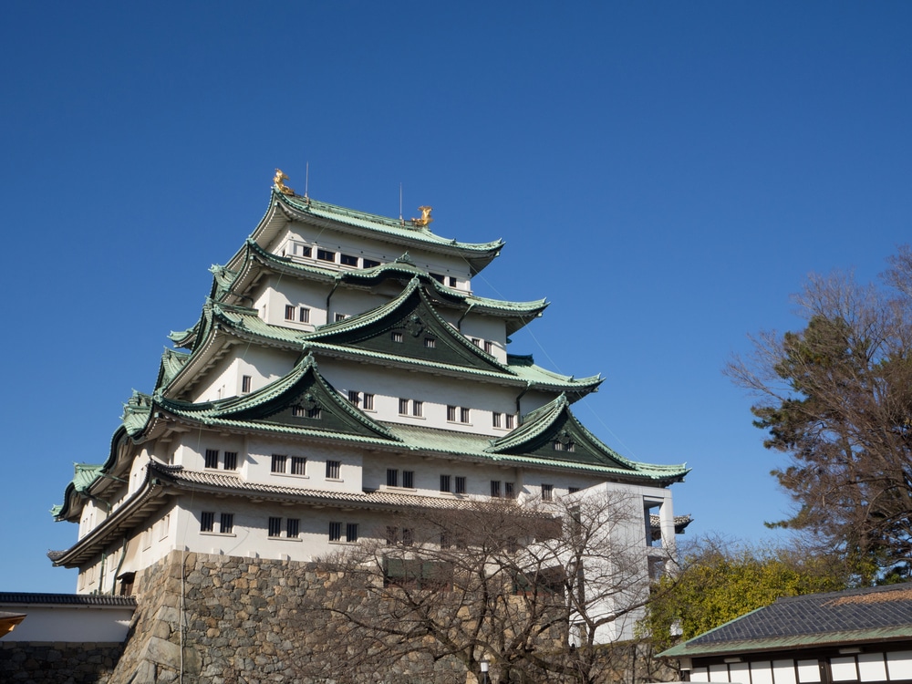 Entdecken Sie Gründe für einen Besuch in Aichi, indem Sie diese traditionelle japanische Burg mit ihren grünen Ziegeldächern und goldenen Details vor einem klaren blauen Himmel erkunden - ein Symbol der verborgenen Mitte Japans.