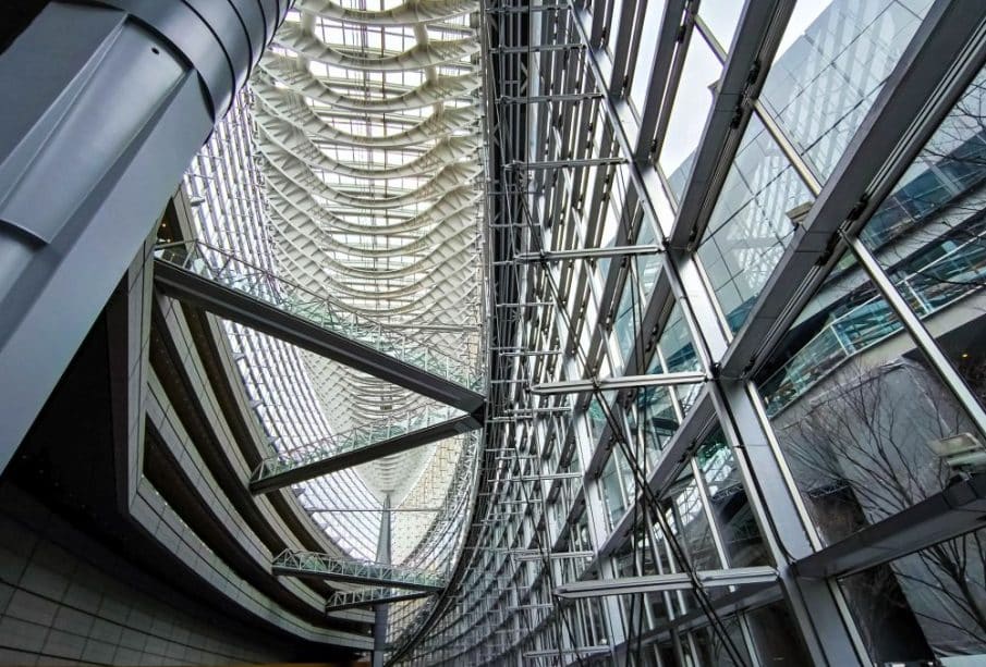 Innenansicht des Tokyo International Forum mit großen Glasfenstern, Metallträgern und einer hohen, geschwungenen Decke mit komplizierter architektonischer Gestaltung. Natürliches Licht durchflutet das geräumige, futuristische Atrium.