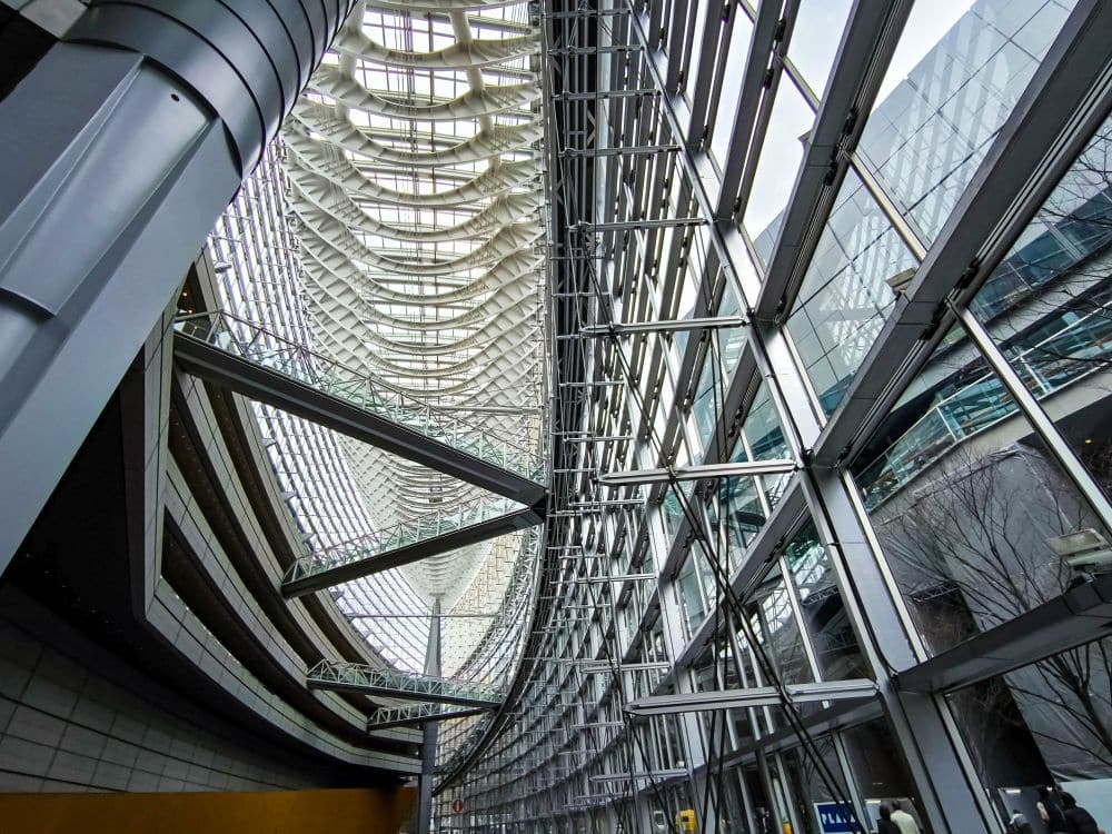 Innenansicht des Tokyo International Forum mit großen Glasfenstern, Metallträgern und einer hohen, geschwungenen Decke mit komplizierter architektonischer Gestaltung. Natürliches Licht durchflutet das geräumige, futuristische Atrium.