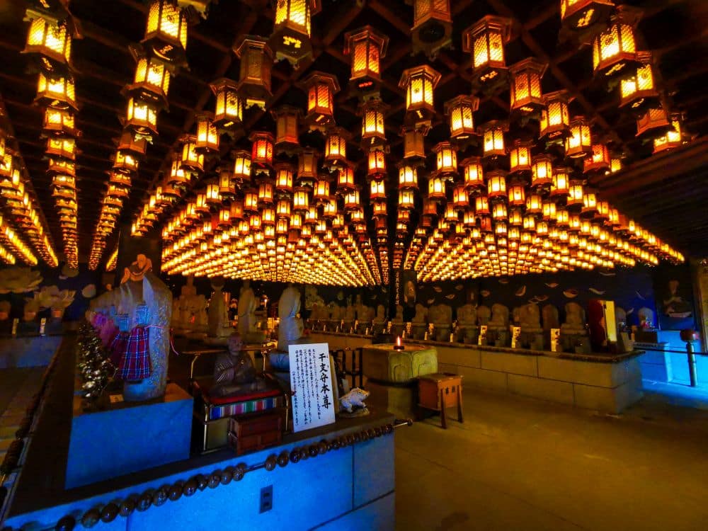 Von der Decke des schwach beleuchteten Daishoin-Tempels hängen Reihen leuchtender Laternen, die die Statuen und Figuren darunter beleuchten und eine warme und heitere Atmosphäre schaffen. Auf einem Schild im Vordergrund ist japanische Schrift zu lesen.