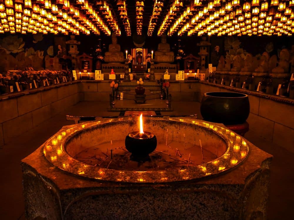 Der hell erleuchtete Innenraum des Daishoin-Tempels mit seinen Reihen leuchtender Laternen an der Decke, einem zentralen Steinaltar mit einer brennenden Kerze sowie Statuen und Opfergaben an den Wänden schafft eine warme und ruhige Atmosphäre.