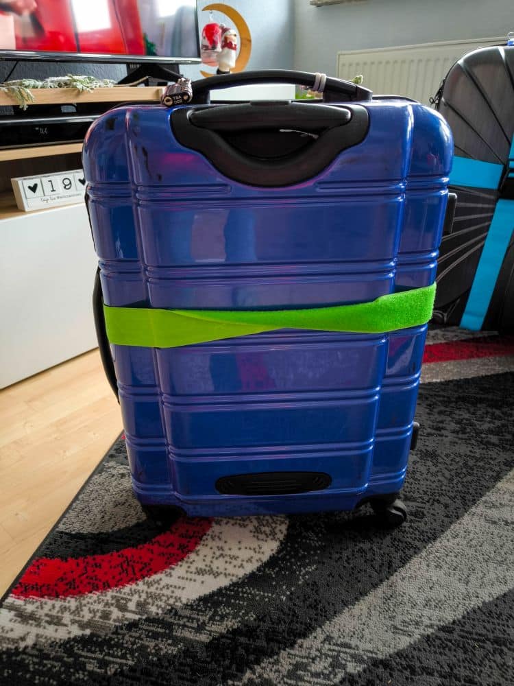 Ein blauer Hartschalenkoffer mit einem hellgrünen Riemen steht auf einem Teppichboden und ist bereit für die Lieferung von Takkyubin. Im Hintergrund befinden sich Regale, ein Heizkörper und ein weiterer Koffer.