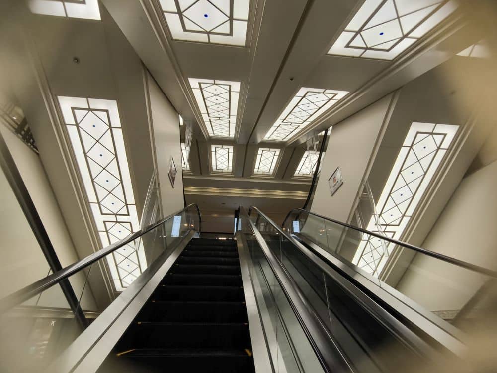 Blick vom unteren Ende einer Rolltreppe, die in einem modernen Gebäude nach oben führt, mit geometrischen Deckenleuchten und hellen, dekorativen Paneelen mit verspielten Anpanman-Motiven an Wänden und Decke.