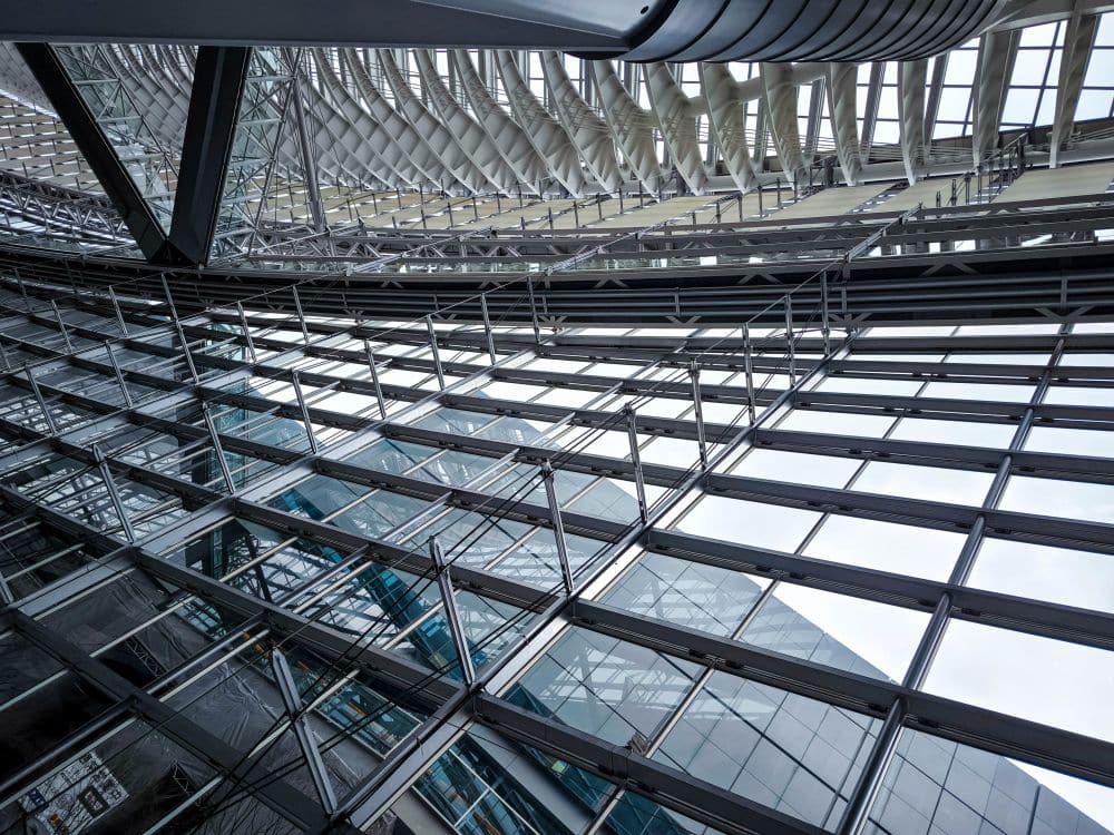 Ein Blick auf den modernen Innenraum des Tokyo International Forum aus Glas und Stahl mit seinen sich kreuzenden Linien, Metallträgern und großen Fenstern mit Spiegelungen, die ein dynamisches geometrisches Muster gegen den Himmel erzeugen.