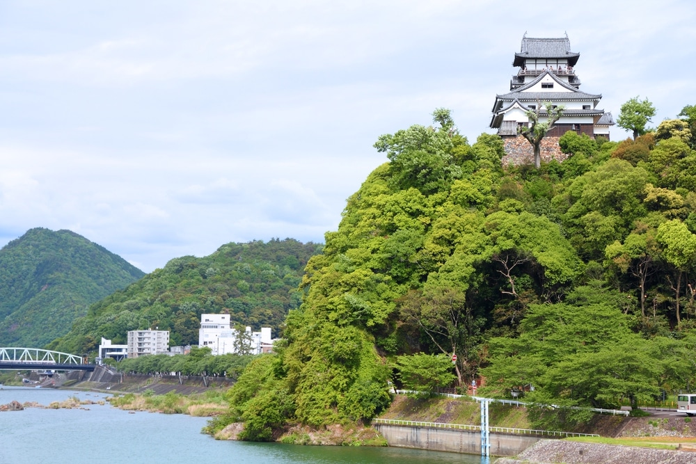 Ein traditionelles japanisches Schloss thront auf einem üppig grünen Hügel in Aichi, Japan, mit Blick auf einen Fluss, mit Bergen im Hintergrund und einigen modernen Gebäuden, die die verborgene Mitte in der Nähe des Wasserrandes zieren.
