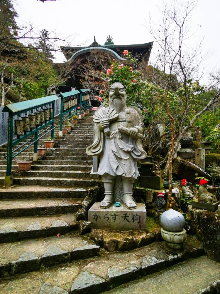 Eine steinerne Statue einer bärtigen Figur, die einen Fächer hält, steht neben einer Steintreppe, die zum Daishoin, einem von Pflanzen und Bäumen umgebenen traditionellen japanischen Tempel, hinaufführt. Am Sockel der Statue sind japanische Schriftzeichen eingraviert.