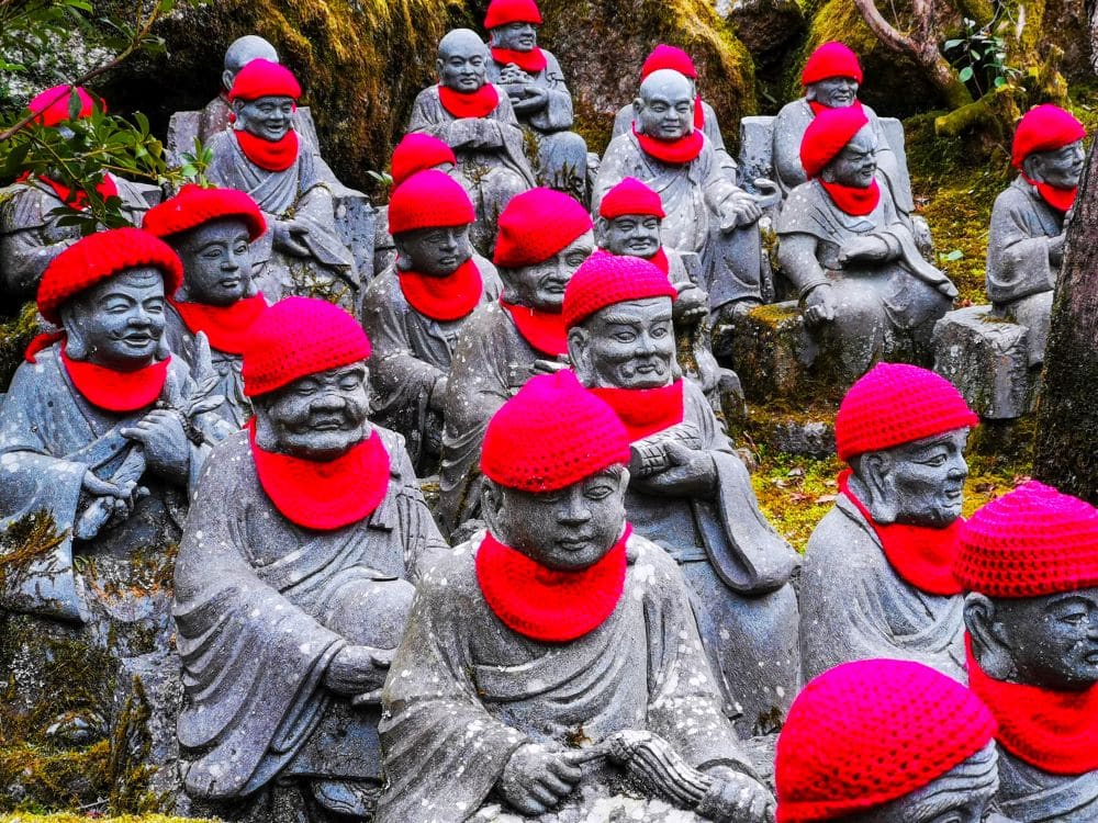 Eine Gruppe steinerner Buddha-Statuen in Daishoin steht im Freien, jede mit einer leuchtend roten Strickmütze und einem Schal geschmückt, umgeben von üppigem Moos und Grün.