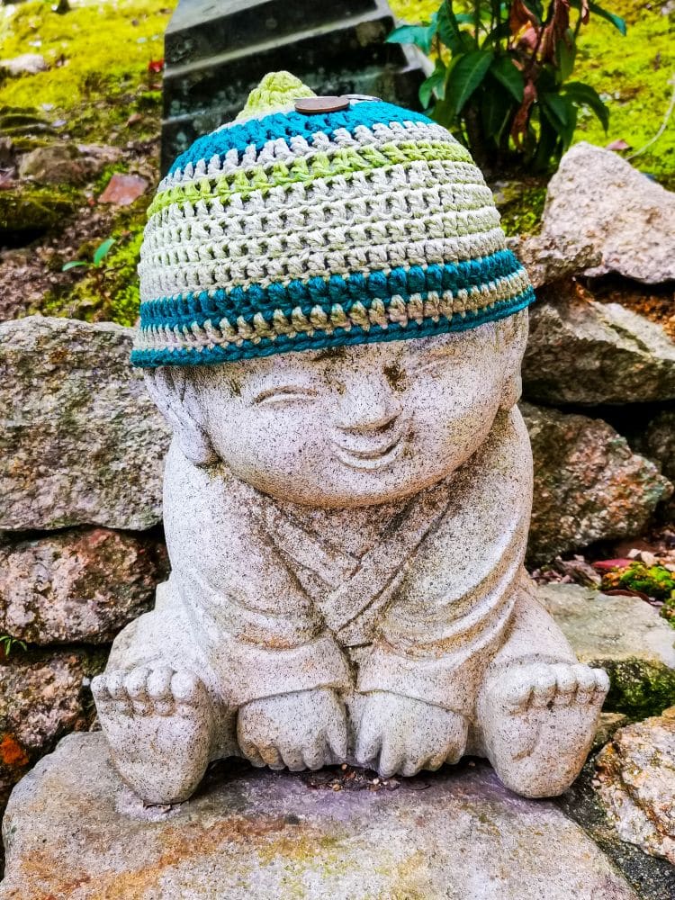 Eine Steinstatue eines lächelnden Kindes sitzt im Schneidersitz zwischen Felsen und Grün im Daishoin-Garten und trägt eine bunte Häkelmütze, die der ruhigen Umgebung Charme verleiht.