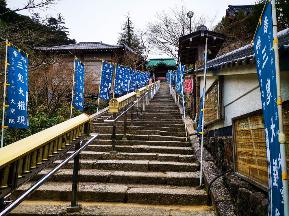 Eine mit blauen vertikalen Bannern gesäumte Steintreppe führt zum Daishoin-Tempel in Japan hinauf, mit Geländern auf beiden Seiten und Bäumen im Hintergrund.
