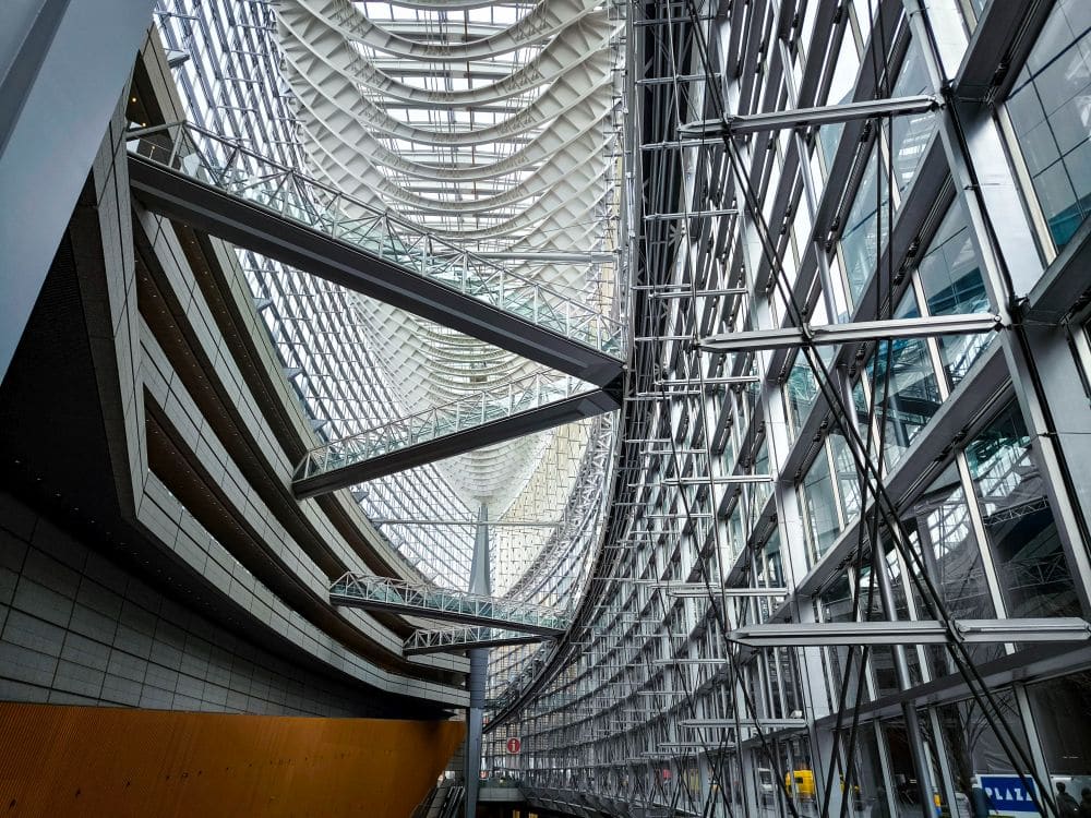 Das moderne architektonische Interieur erinnert an das Tokyo International Forum, mit gebogenen Stahlträgern, großen Glasflächen und natürlichem Licht, das eine großzügige, futuristische Atmosphäre schafft. Strukturelle Elemente und Gehwege sind überall im Raum sichtbar.