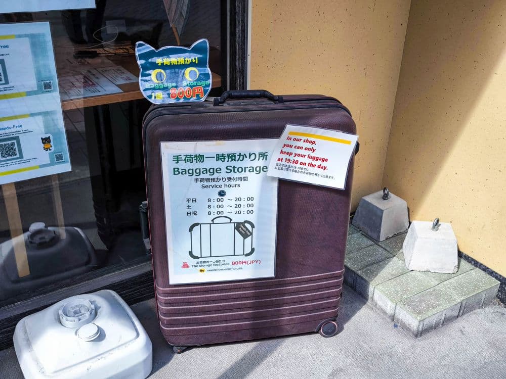 Ein großer brauner Koffer steht vor einem Gebäude mit einem Schild in Japanisch und Englisch, das für die Gepäckaufbewahrung und den Takkyubin-Service wirbt und die Servicezeiten angibt. Ein weiteres Schild weist darauf hin, dass der Preis für die Aufbewahrung 800 Yen pro Tag beträgt.