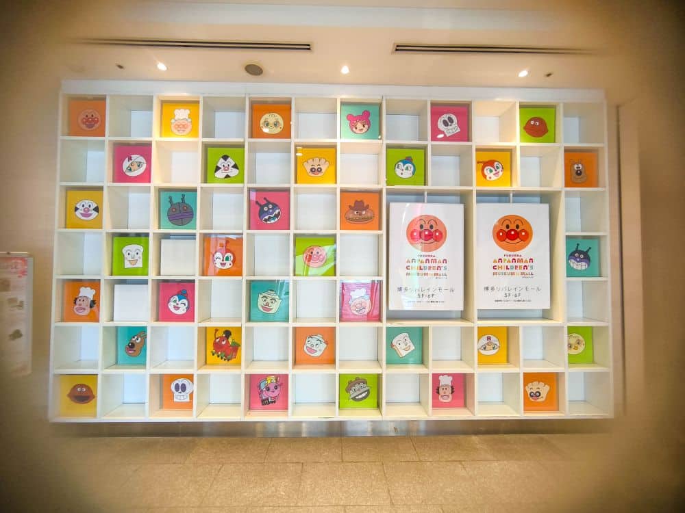 An einer farbenfrohen Wand sind verspielte Anpanman-Cartoonfiguren in quadratischen, teilweise leeren Fächern zu sehen. Zwei Plakate in der Mitte zeigen japanische Texte und den Schriftzug "Anpanman Children's Museum", die zu der fröhlichen Szene beitragen.