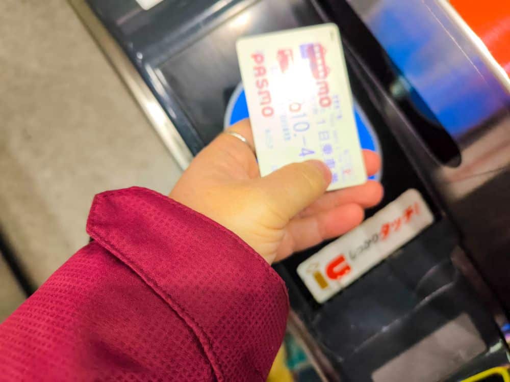 Eine Person in einer roten Jacke hält eine PASMO-Karte über ein Metro-Kartenlesegerät an einem Fahrkartenschalter in einem japanischen Bahnhof. Das Foto ist von oben aufgenommen und zeigt, wie die Karte für den Eintritt gescannt wird.
