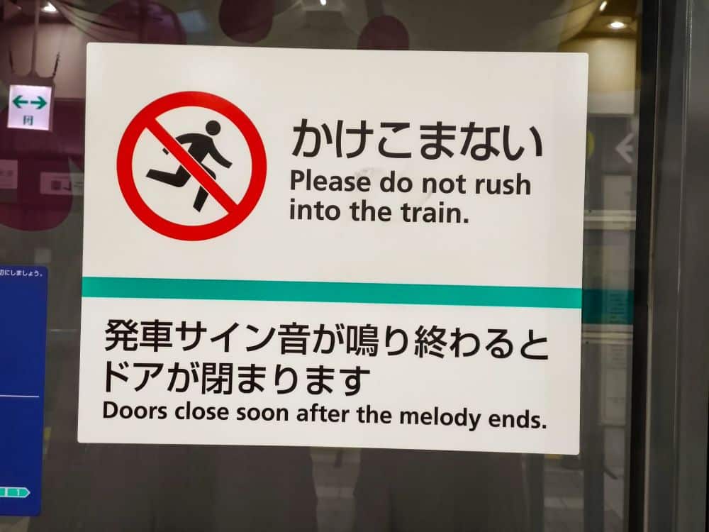 Auf einem zweisprachigen Metro-Schild am Bahnhof steht: "Bitte drängen Sie nicht in den Zug" und "Die Türen schließen kurz nach Ende der Melodie". Es zeigt ein rotes Verbotszeichen mit einer rennenden Person, sowohl auf Japanisch als auch auf Englisch.