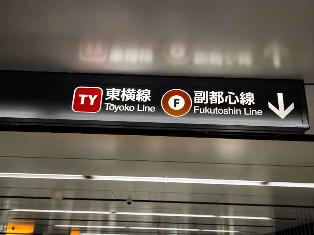 Ein Metro-Schild zeigt die Richtungen für die Toyoko-Linie (TY) und die Fukutoshin-Linie (F) in Englisch und Japanisch an, mit farbigen Symbolen und einem weißen Pfeil, der nach unten zeigt. Das Schild ist an einer Innendecke angebracht.