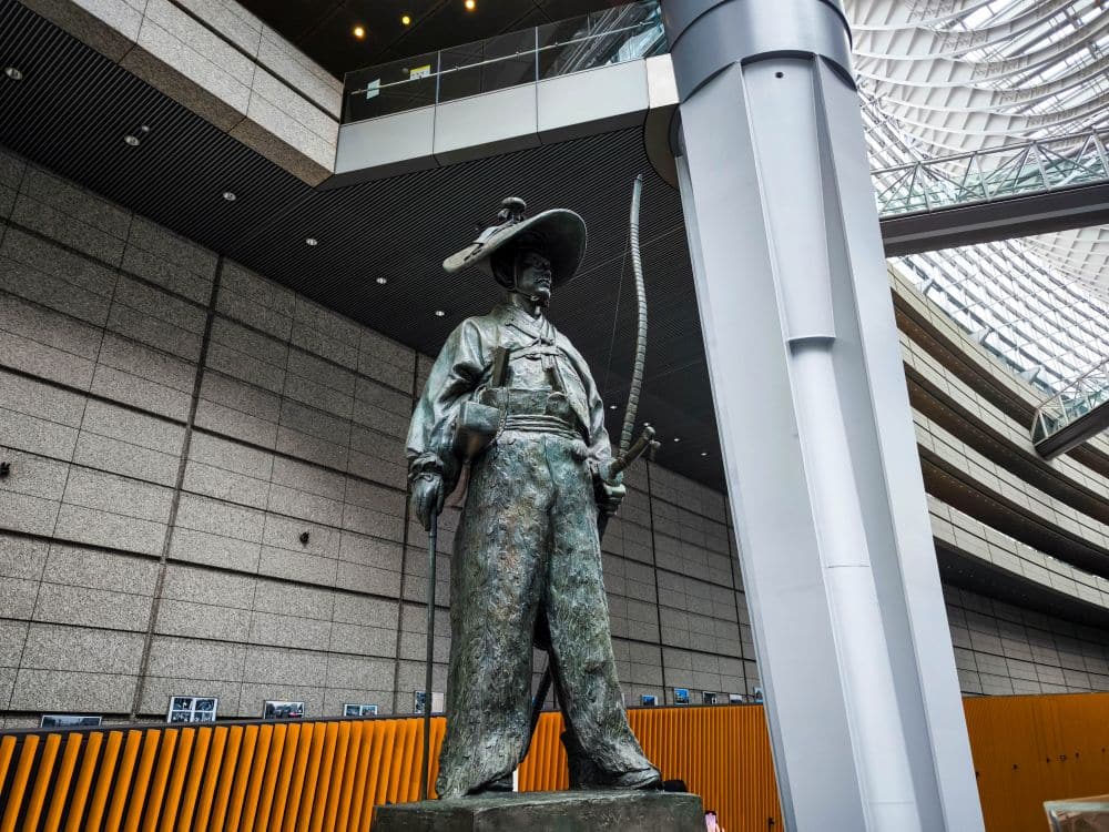 Eine Bronzestatue eines Samurai in traditioneller Kleidung steht im Innenbereich des Tokyo International Forum, hält einen langen Bogen und trägt einen Hut. Im Hintergrund sind moderne architektonische Elemente und eine Glasdecke zu sehen.