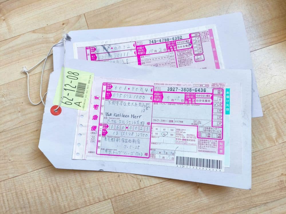 Zwei japanische Takkyubin-Versandetiketten mit handschriftlichen Angaben und Strichcodes sind an weißen Anhängern befestigt, die auf einem hellen Holzboden liegen. Ein Etikett hat einen gelben Aufkleber mit der Aufschrift 62-12-08 und einen roten Ring in der Ecke.