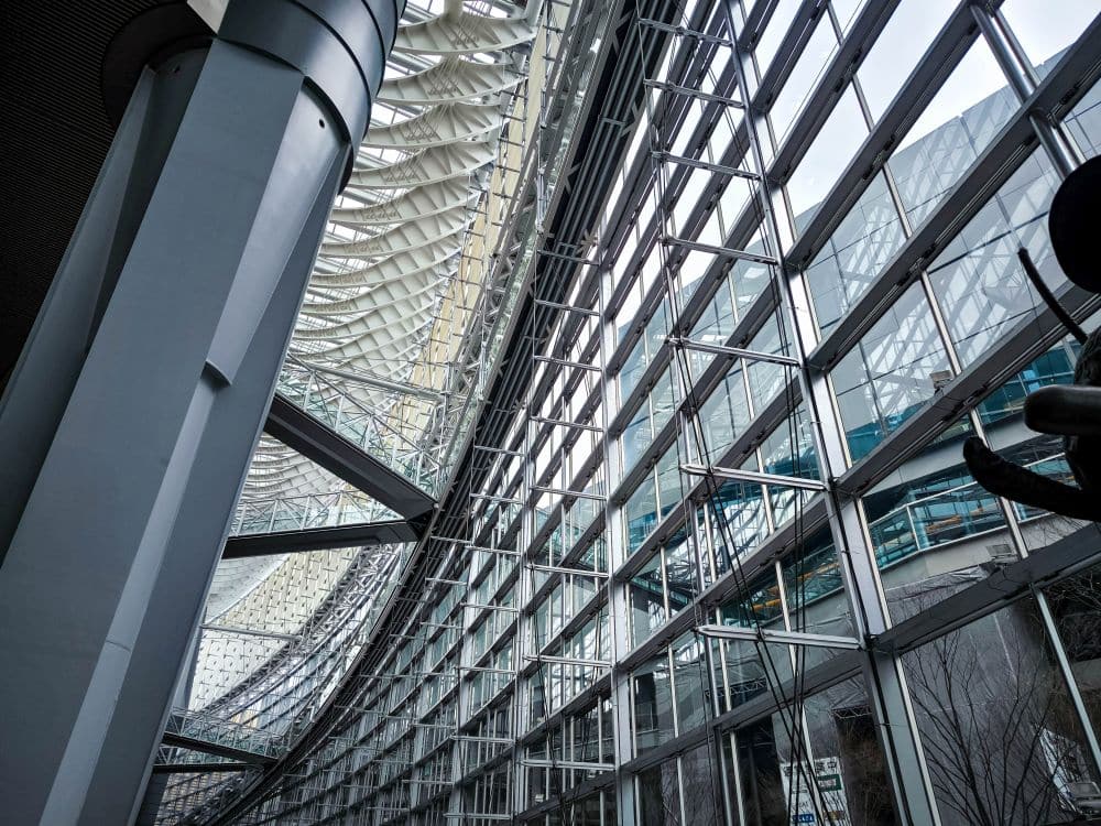 Das moderne architektonische Interieur mit einer hohen, geschwungenen Wand aus Glas und Stahl, die an das Tokyo International Forum erinnert, weist strukturelle Balken und geometrische Deckenmuster auf. Durch die großen Fenster strömt natürliches Licht, das Reflexionen und ein Gefühl von Großzügigkeit erzeugt.