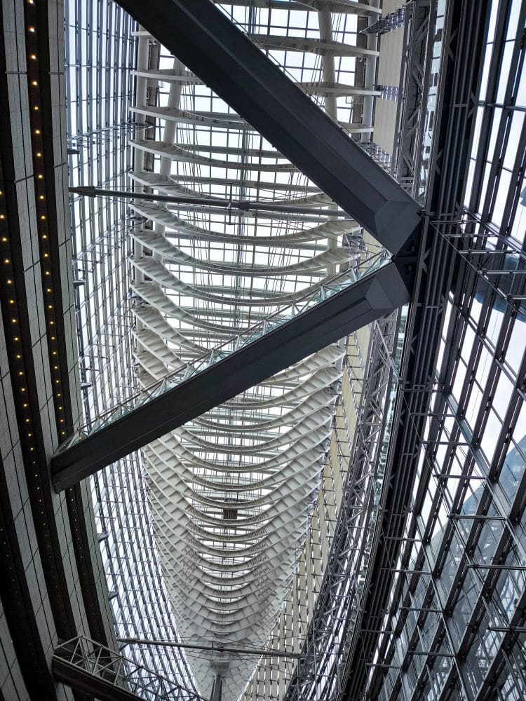 Innenansicht des Tokyo International Forum mit einer hohen, gebogenen Glasdecke und freiliegenden, sich kreuzenden Metallträgern. Das architektonische Design zeichnet sich durch sich wiederholende geometrische Muster und schlanke, moderne Linien aus, die in natürliches Licht getaucht sind.