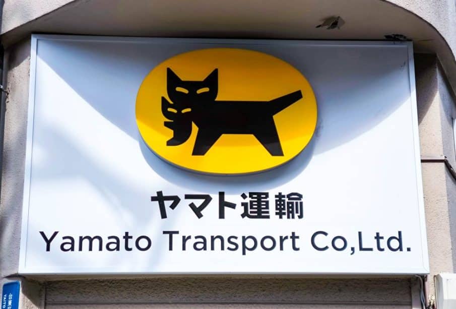 Ein Schild für Yamato Transport Co., Ltd. zeigt eine schwarze Katze, die ein Kätzchen im Maul trägt, auf einem gelben ovalen Hintergrund, darunter Takkyubin und den Firmennamen in Japanisch und Englisch.