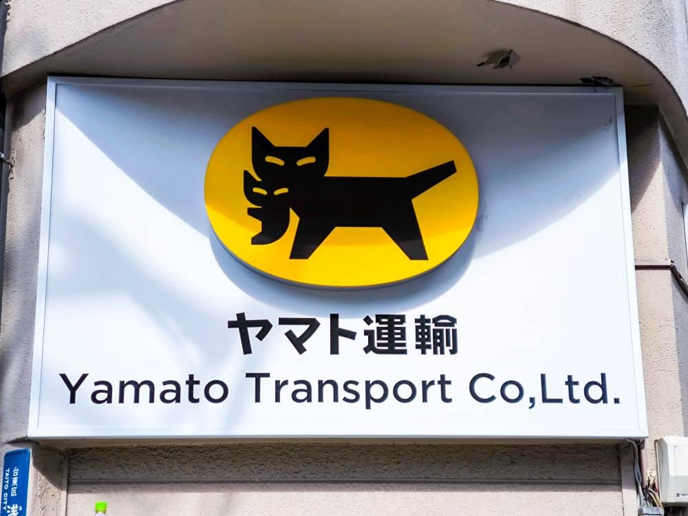 Ein Schild für Yamato Transport Co., Ltd. zeigt eine schwarze Katze, die ein Kätzchen im Maul trägt, auf einem gelben ovalen Hintergrund, darunter Takkyubin und den Firmennamen in Japanisch und Englisch.