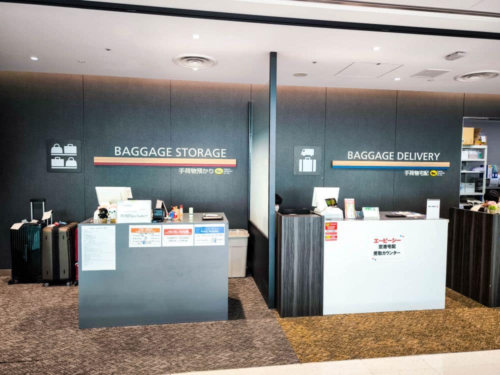Zwei Schalter mit der Aufschrift Baggage Storage und Takkyubin Baggage Delivery stehen nebeneinander, mit Schildern in Englisch und Japanisch. Koffer, Computer und Papierkram sind an den Schaltern in einer sauberen, modernen Umgebung zu sehen.