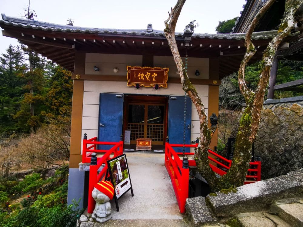 Ein kleiner japanischer Tempel mit rotem Geländer, einem Ziegeldach und einem Holzschild über dem Eingang, umgeben von Bäumen und Grünpflanzen. Eine Treppe führt hinauf zum Daishoin-Tempel. Im Vordergrund ist eine Steinlaterne zu sehen.