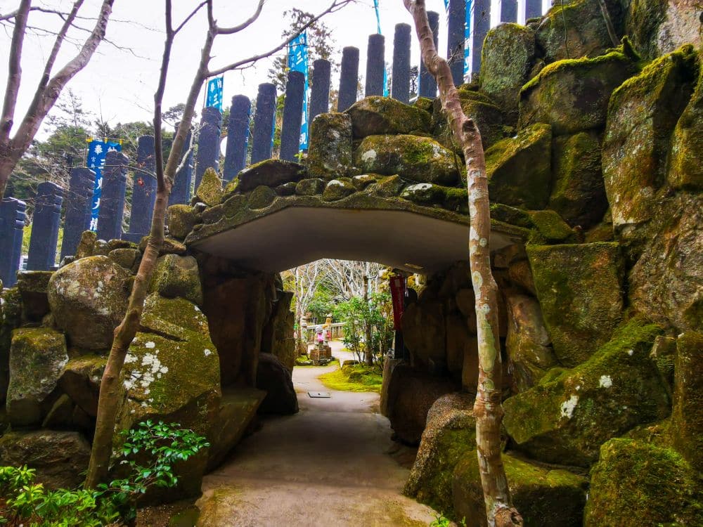 Ein steinerner Torbogen, der in große Felsen eingelassen ist, führt zu einem mit Bäumen und Grünpflanzen gesäumten Gartenweg, über dem hohe Steinsäulen stehen, die an die ruhige Schönheit von Daishoin erinnern.