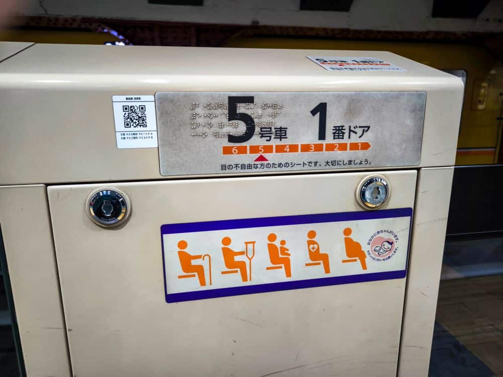 Ein Schild auf einem Bahnsteig der U-Bahn weist auf reservierte Sitzplätze für Behinderte, Verletzte, Schwangere oder Personen mit Kindern hin. Das Schild besteht aus japanischem Text, Zahlen und Symbolen, die die vorrangigen Sitzplatzkategorien darstellen.