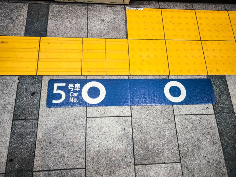 Ein U-Bahn-Bahnsteig ist mit taktilen gelben Kacheln für Sehbehinderte und einem blauen Schild mit der Aufschrift 5 Car No. auf Englisch und Japanisch versehen, das den Einstiegsort für den U-Bahn-Wagen 5 angibt.
