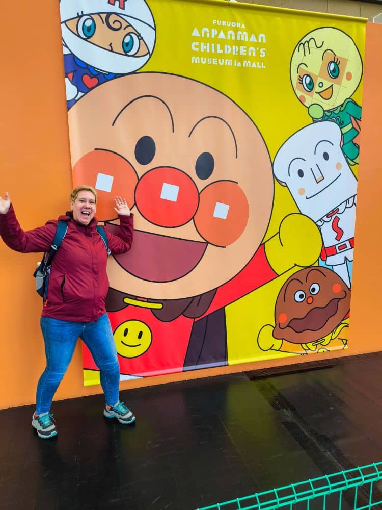 Eine Person in einer kastanienbraunen Jacke lächelt aufgeregt vor einem farbenfrohen Anpanman-Poster im Anpanman-Kindermuseum in Fukuoka, das große Zeichentrickfiguren auf einem leuchtend gelben und orangen Hintergrund zeigt.