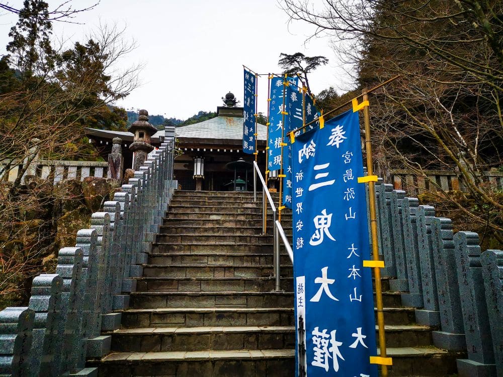 Steinstufen führen hinauf zum Daishoin, einem traditionellen japanischen Schrein, der von blauen vertikalen Bannern mit weißen Kanji-Schriftzeichen flankiert wird und von Bäumen und Bergen im Hintergrund umgeben ist.