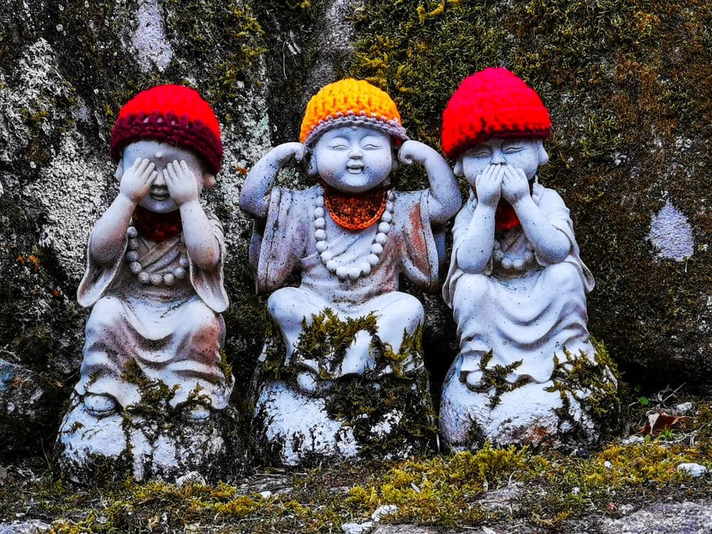 Drei Steinstatuen von Mönchen in Daishoin sitzen Seite an Seite, jeder mit einer bunten Strickmütze und einer Halskette. Sie stehen für "Sieh nichts Böses, hör nichts Böses, sprich nichts Böses" und ruhen mit bedeckten Augen, Ohren und Mund zwischen moosbewachsenen Felsen.